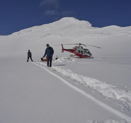 Two people standing in the snow next to a helicopter on top of a mountain.