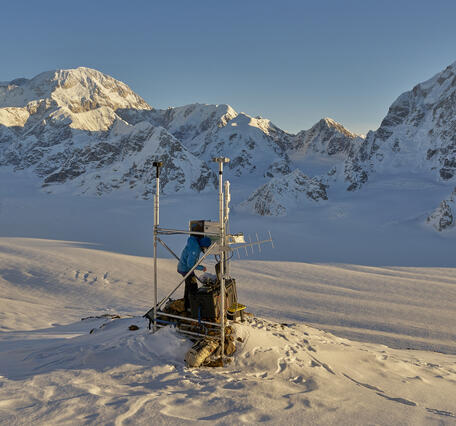 Person in a blue jacket working at a weather station in the snow with mountains in the background 
