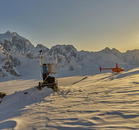 Person in blue jacket at a weather station on snow with the sunshine, mountains, and a red helicopter in the background