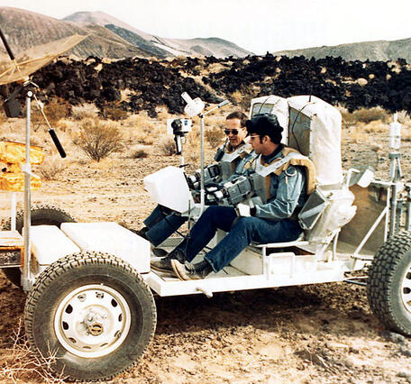 Two white men wearing jeans, button-down shirts, and bulky harnesses with huge rectangular backpacks sit on the frame of a stripped-down white motorized buggy-like vehicle. Their harnesses have two old-fashioned video cameras pointing forward. On the front of the vehicle, a large metallic mesh radio antenna dish is raised on a pole. In the background, mounds of dark, jagged, black lava rock sit in front of a skyline composed of several rounded red hills of volcanic scoria. 