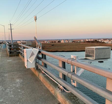 Looking along a bridge rail where a radar unit is suspended out over the river and tied into a control box with a rain gage