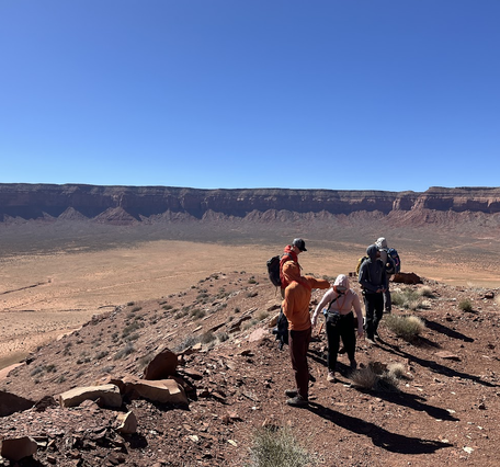 Five people stand in the Utah desert viewing the rocks around them with a large cliff in the background.