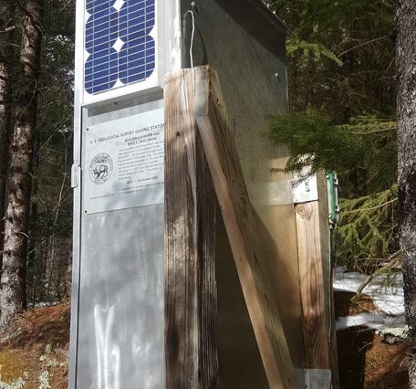 Gray metal USGS streamgage housing with solar panel and inscription mounted on a wooden support in a forested area.
