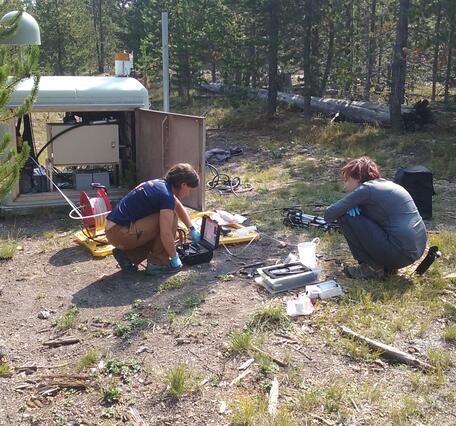 Two people crouching near equipment in a forested clearing and working with hand tools