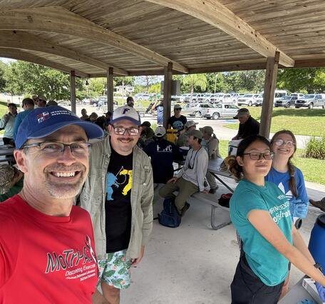 Four people stand in the foreground taking a selfie-style photo, with many people sitting on metal park benches under a large pavilion in the mid-ground, and a road and parking lot in the background