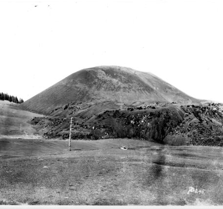 A historic black-and-white photo shows a small, round mountain made up of barren rocks. A huge lava flow spreads from its base, sparsely dotted by spindly pine trees. 