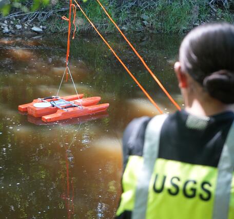 An acoustic Doppler current profiler is in focus with a person wearing a USGS personal flotation device in the foreground