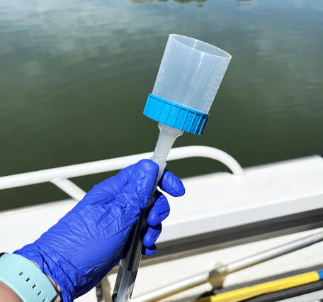 A hand with a blue glove on holds an eDNA sampling filter next to the edge of a boat in a reservoir