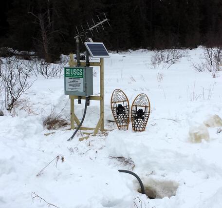 A pair of snowshoes rests in the snow next to a small streamgage mounted on a wooden frame, with antenna and solar panel.