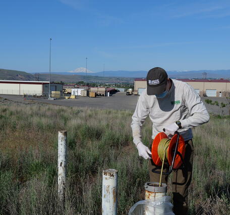 Scientist lowers tape into well in a field with warehouse