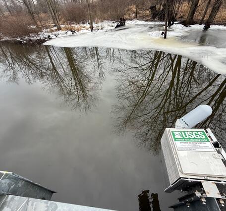 A streamgage housing is mounted on the side of a bridge over a creek. Ice covers the shoreline.