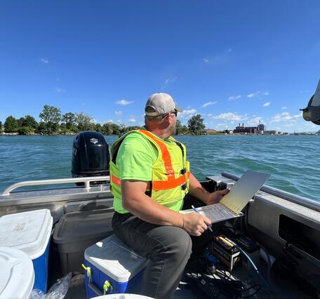 Hydrologic technician on a boat, holding a laptop looks over a blue river