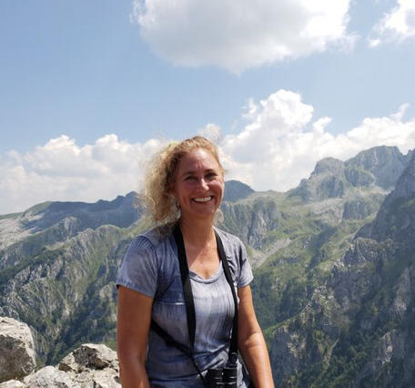 A woman with blonde hair smiles towards the camera with mountains behind her