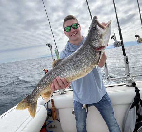 A person on a boat holds up a large fish with speckled coloring and a golden tail. Several fishing rods are positioned upright at the back of the boat, and open water extends into the distance beneath a cloudy sky. 