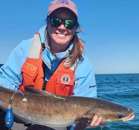 A peson in a hat and sunglasses and orange life jacket leans on the side of a boat holding a cobia