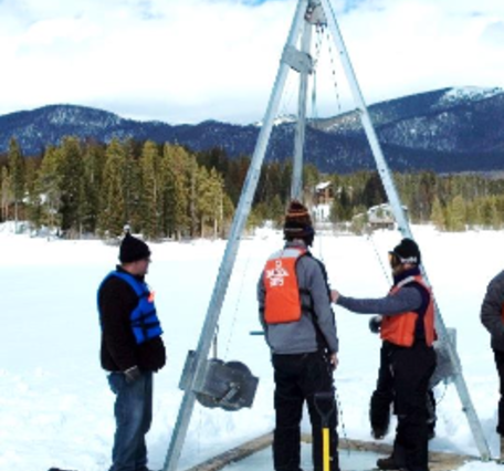 scientists in winter clothes and safety gear examine a percussion piston coring system situated on a frozen lake