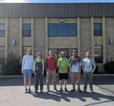 Six people pose in front of a brown brick building on a sunny summer day