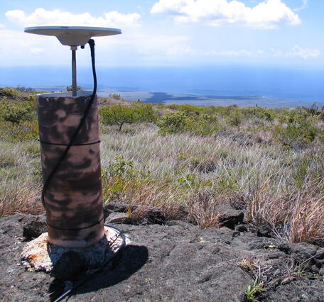 Color photograph of volcano monitoring device with grass in the foreground and ocean in the background
