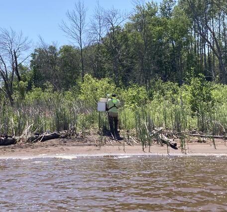 Hydrologic technician stands on shoreline and installs equipment in a streamgage housing