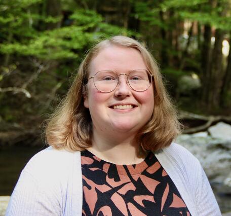 A woman with shoulder length blonde hair wearing a pink and black blouse with a white jacket smiles for her picture. 