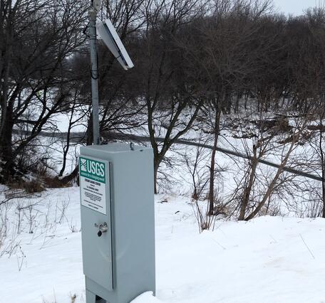USGS streamgage with solar panel beside a snow-covered riverbank and leafless trees in winter