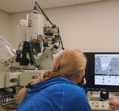 Blonde scientist in a blue hoodie sitting in front of a electron microprobe and computer screen showing a black-and-white image of a crystal
