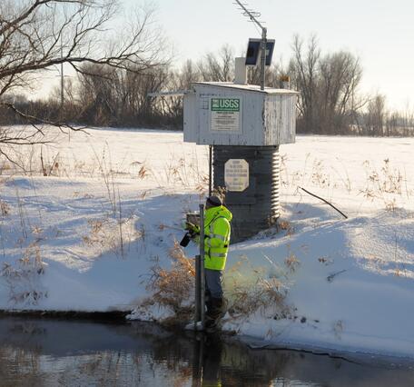 A hydrologic technician wearing a bright yellow coat stands in front of a streamgage housing and takes a streamflow reading