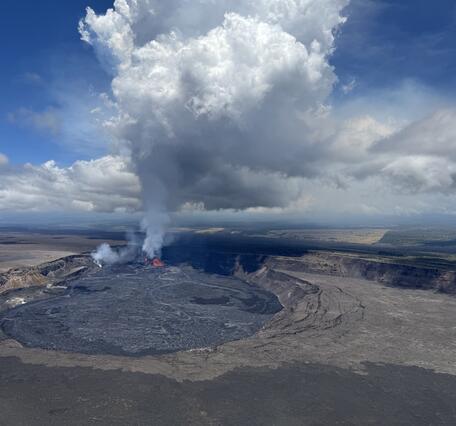 A large white and blue plume rises from the red lava fountains within a crater