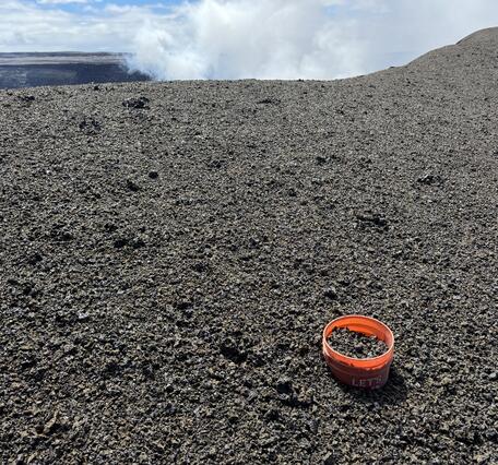 Color photograph of volcanic terrain with bucket in foreground and degassing in background
