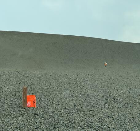 Color photograph of bucket next to mound of volcanic material with person walking in the distance