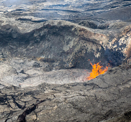 Color photo of bright red lava emerging from lava lake with cooled, black surface. 