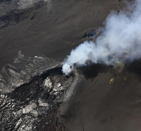 color photo looking down at cooled black lava and pale gray steam.