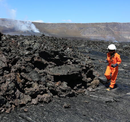 A scientists wearing and orange flight suit and a white flight helmet samples black, cooled lava. 