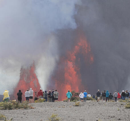 Color photograph of people standing on the rim of a crater with lava fountains in the background