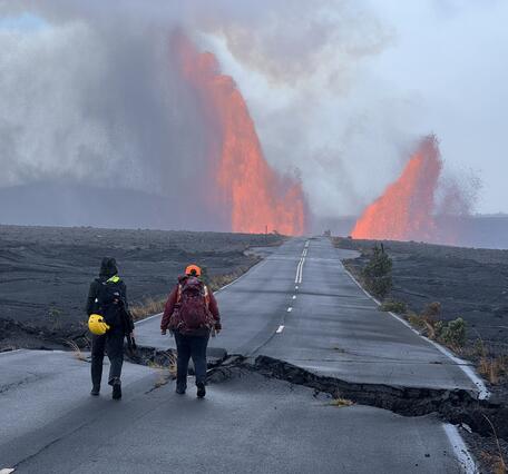 Color photograph of scientists walking on damaged road with lava fountains in the background