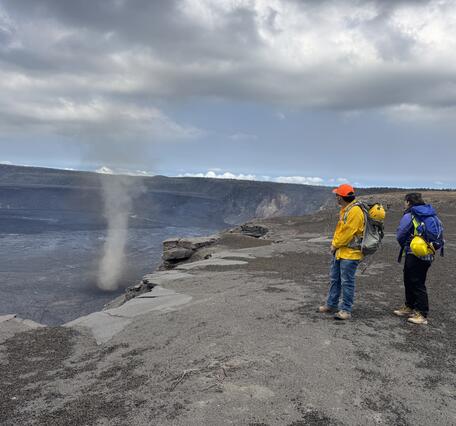 Color photograph of scientists on the edge of a volcanic crater with a whirlwind in it