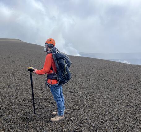 Color photograph of scientist wearing personal protective equipment and taking a measurement on a crater rim