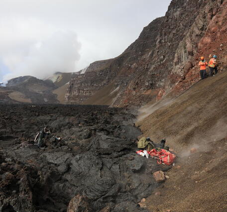 Color photograph of scientists sampling crater wall with volcanic vents degassing in background