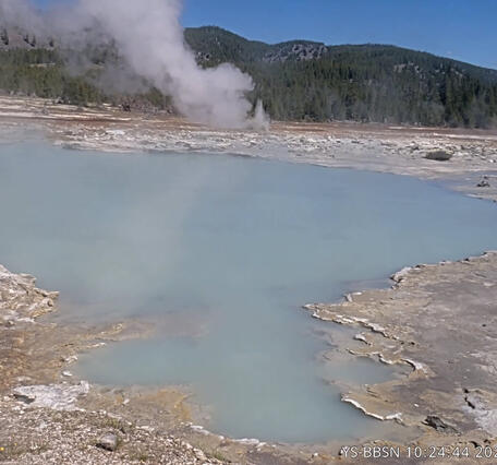 blue pool in a barren area with a treed ridge in the background under blue sky and a small steam vent in the middle ground