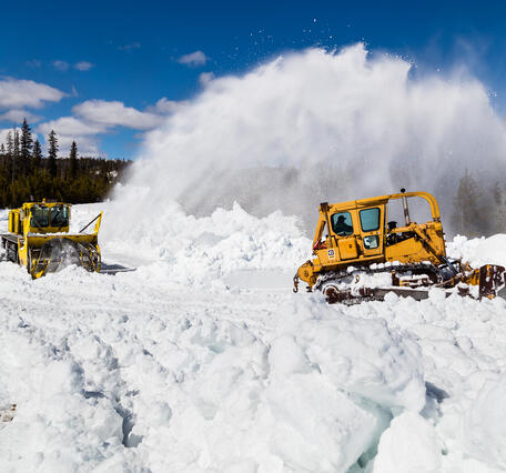 Snowplow removing snow form a road, with trees in the background under partly cloudy skies