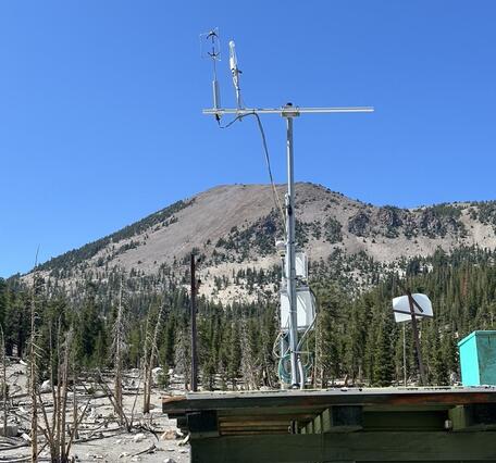A tall white metal tower with scientific instruments stands on a rooftop of a small building in a mountainous area with scattered rocks and sparse vegetation. In the background, Mammoth Mountain rises as a forested peak under a clear blue sky. The monitoring equipment includes sensors mounted at the top of a 6-meter-high mast for measuring volcanic CO₂ emissions.