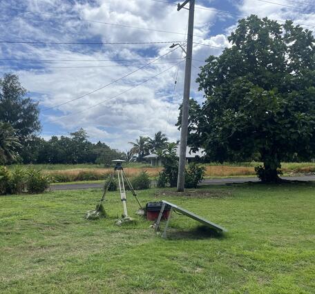 Color photo of a GPS station and solar panel in grass in a tropical setting.