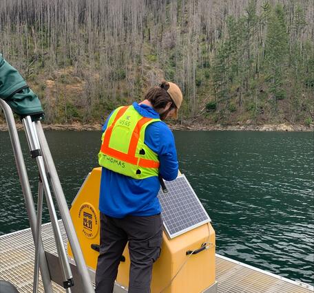 male uses a tool to open gear box on a floating platform in green lake water on a cloudy day. Burned trees in background