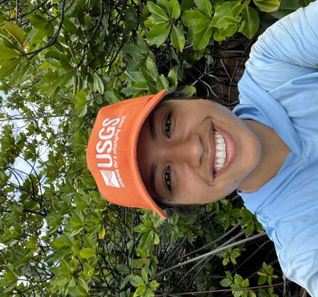 Photo of person wearing a light blue shirt and an orange USGS hat in front of vegetation.