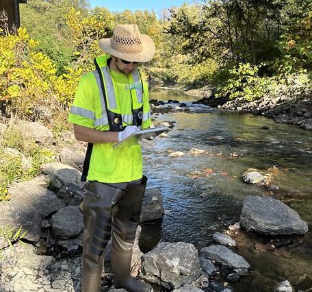 Scientist records conditions of river while standing next to it with a highway overpass in background