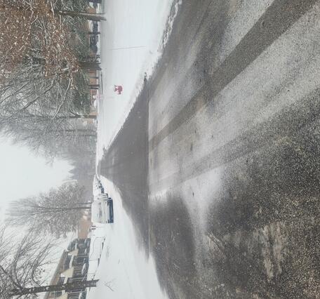 Road in neighborhood covered in snow with heavy salt on ground. Car tire tracks going through salt. 