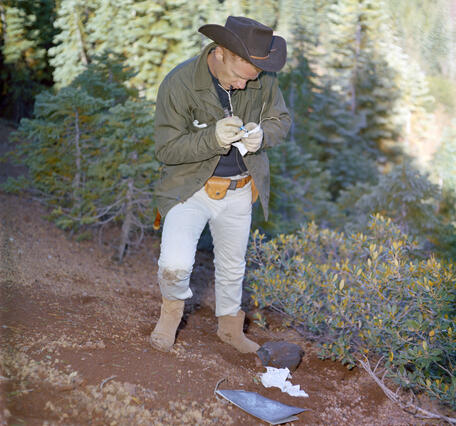 A red-haired man wearing khaki pants, brown boots, a green jacket, and a black cowboy hat stands on a steep, reddish slope surrounded by manzanita bushes and small pine trees. He is taking notes in a small notebook. In the background, a thick pine forest is lit by sunlight.