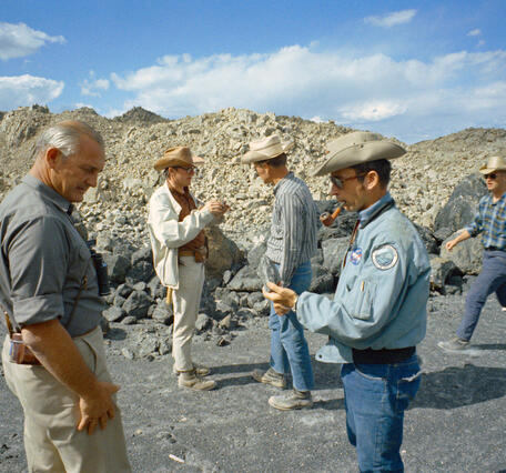 A group of five white men wearing cowboy hats, jeans or khakis, and button-down shirts are standing on a sandy gray surface ringed by large gray boulders of volcanic rock. In the background, a number of bare, rubbly, rocky peaks are lit by afternoon sun. The men are examining hand samples of rocks and one is smoking a pipe.