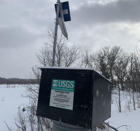 USGS streamgage housing with solar panel and sensors mounted on a pole, against a snow-covered rural background