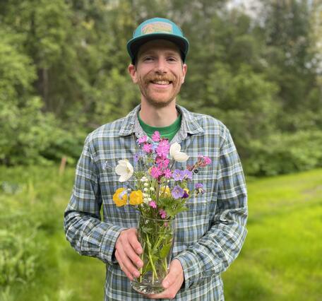 A man wearing a plaid button up and a baseball cap holds a bouquet of flowers.